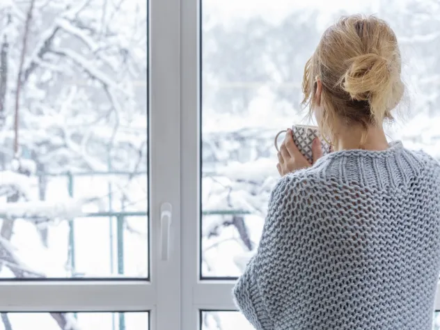 Kvinna med tekopp tittar ut genom fönstret mot ett snöigt landskap.