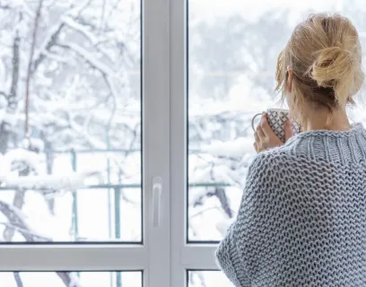 Kvinna med tekopp tittar ut genom fönstret mot ett snöigt landskap.