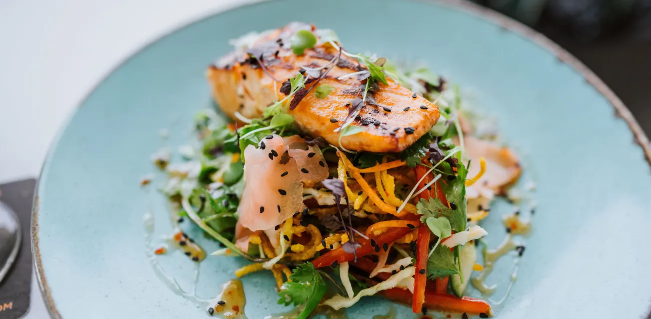 Gourmettallrik med grillad lax, inlagda och färska grönsaker och topping på en ljusblå tallrik.