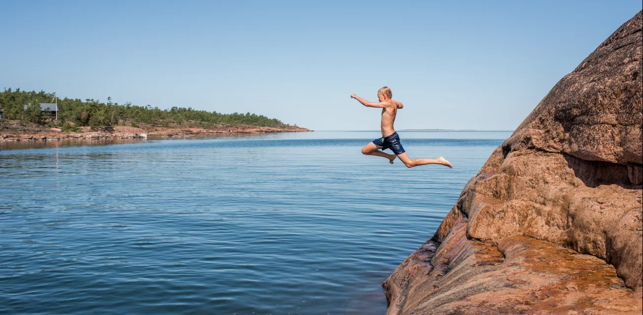 Pojke hoppar i vattnet från badklippa på Åland.