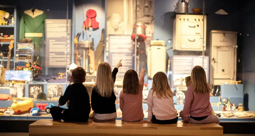 Ryggarna på barn som sitter på en bänk och pekar på olika saker i basutställningen vid Ålands kulturhistoriska museum.